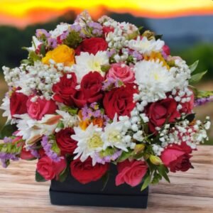 A stunning floral arrangement featuring red and pink roses, white chrysanthemums, baby’s breath, and touches of yellow and purple blooms, all elegantly presented in a black box. The vibrant colors create a beautiful contrast, making it a perfect choice for a heartfelt gift or a decorative centerpiece. The breathtaking sunset in the background adds warmth and romance to the scene.