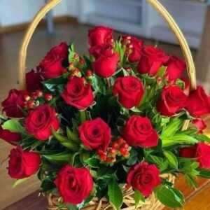 Basket arrangement of red roses with green foliage and red hypericum berries placed on a wooden surface.