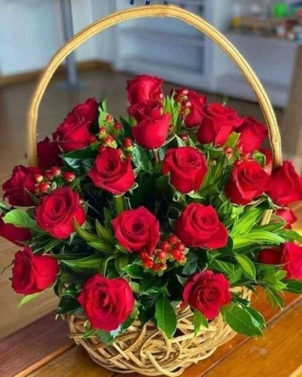 Basket arrangement of red roses with green foliage and red hypericum berries placed on a wooden surface.