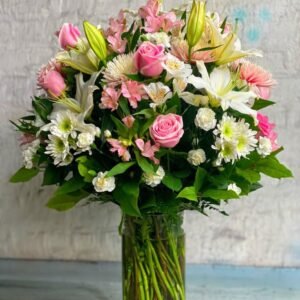 Bouquet of pink roses, white lilies, and assorted flowers with green foliage in a clear glass vase.