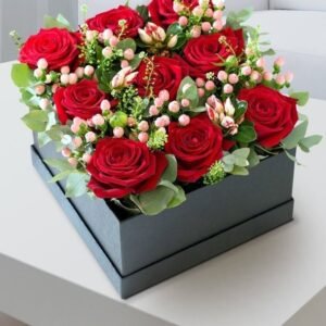 Box arrangement of red roses, pink berries, and greenery in a black gift box.