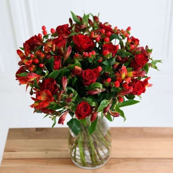 Bouquet of red roses and alstroemeria with green foliage arranged in a clear glass vase on a wooden surface.