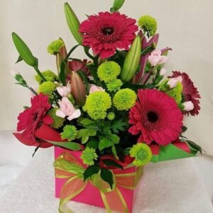 Floral arrangement in a pink gift box with a green ribbon, featuring red gerbera daisies, green chrysanthemums, pink lilies, and assorted greenery.