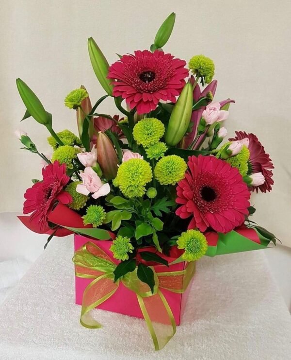 Floral arrangement in a pink gift box with a green ribbon, featuring red gerbera daisies, green chrysanthemums, pink lilies, and assorted greenery.