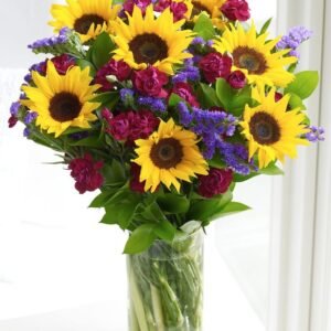 Vibrant bouquet of sunflowers, red roses, and purple statice arranged in a clear glass vase with lush greenery