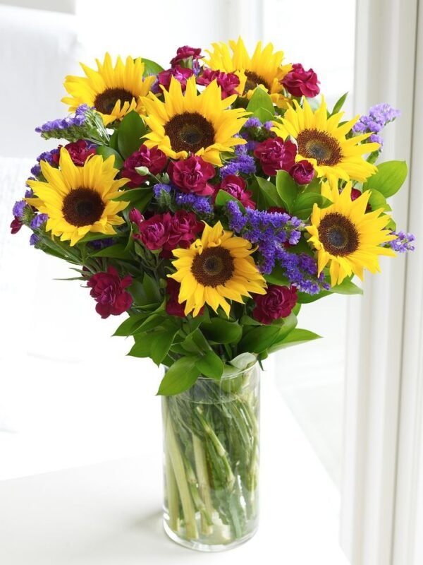 Vibrant bouquet of sunflowers, red roses, and purple statice arranged in a clear glass vase with lush greenery