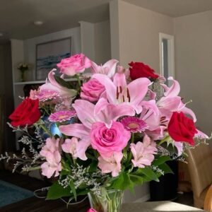 Beautiful bouquet of pink lilies, pink and red roses, gerbera daisies, and lush greenery arranged in a glass vase.