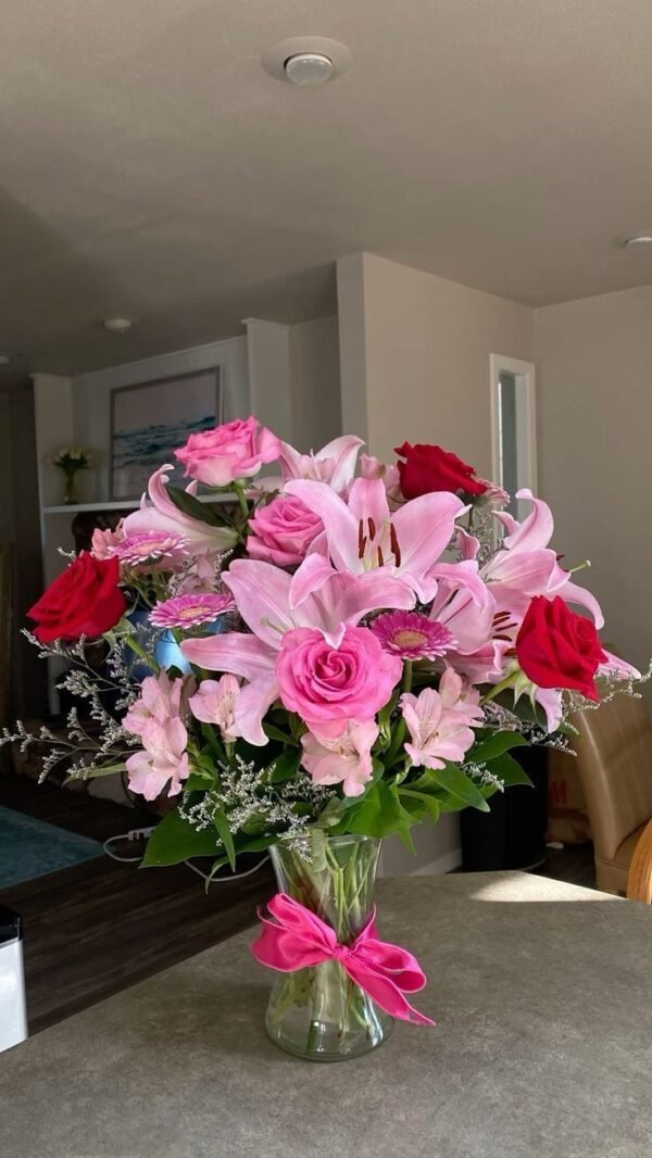 Beautiful bouquet of pink lilies, pink and red roses, gerbera daisies, and lush greenery arranged in a glass vase.