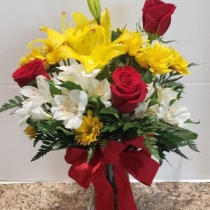 Elegant floral arrangement featuring red roses, yellow lilies, white alstroemeria, and yellow daisies, accented with greenery and a red ribbon, displayed in a clear vase on a marble countertop