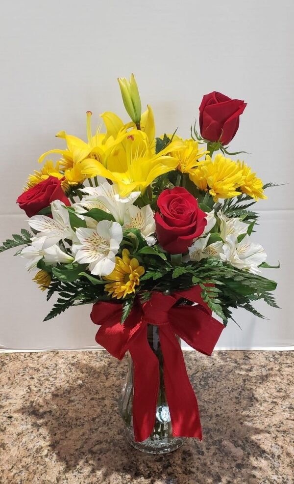 Elegant floral arrangement featuring red roses, yellow lilies, white alstroemeria, and yellow daisies, accented with greenery and a red ribbon, displayed in a clear vase on a marble countertop