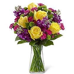 Elegant floral arrangement featuring yellow roses, purple flowers, and green accents in a clear glass vase.