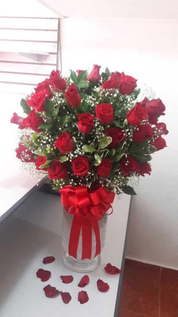 Luxurious bouquet of red roses with baby's breath and lush greenery, arranged in a glass vase with a red ribbon.